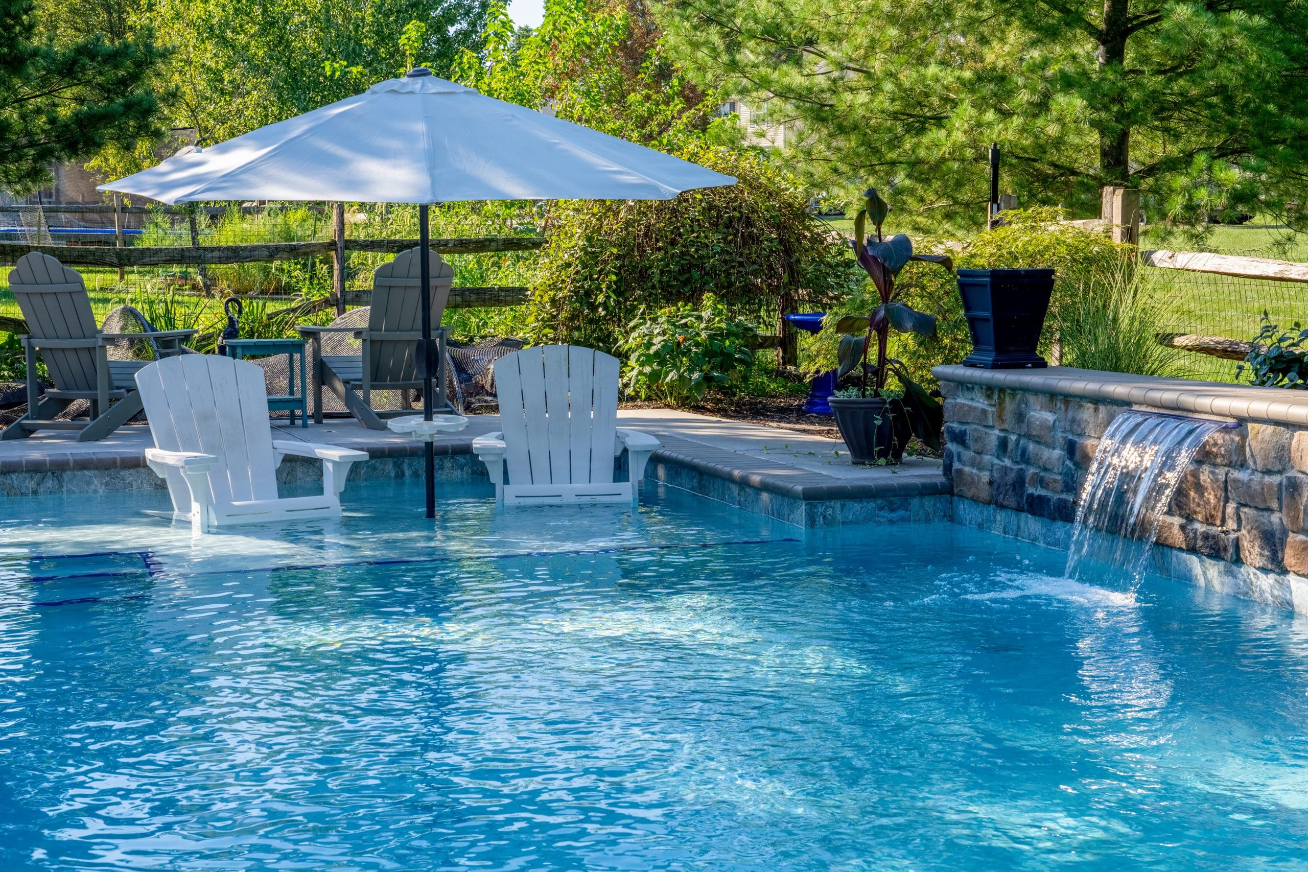 A backyard pool with clear blue water, two white chairs partly submerged under a large umbrella, a stone waterfall feature, patio seating, lush greenery, and a wooden fence in the background.