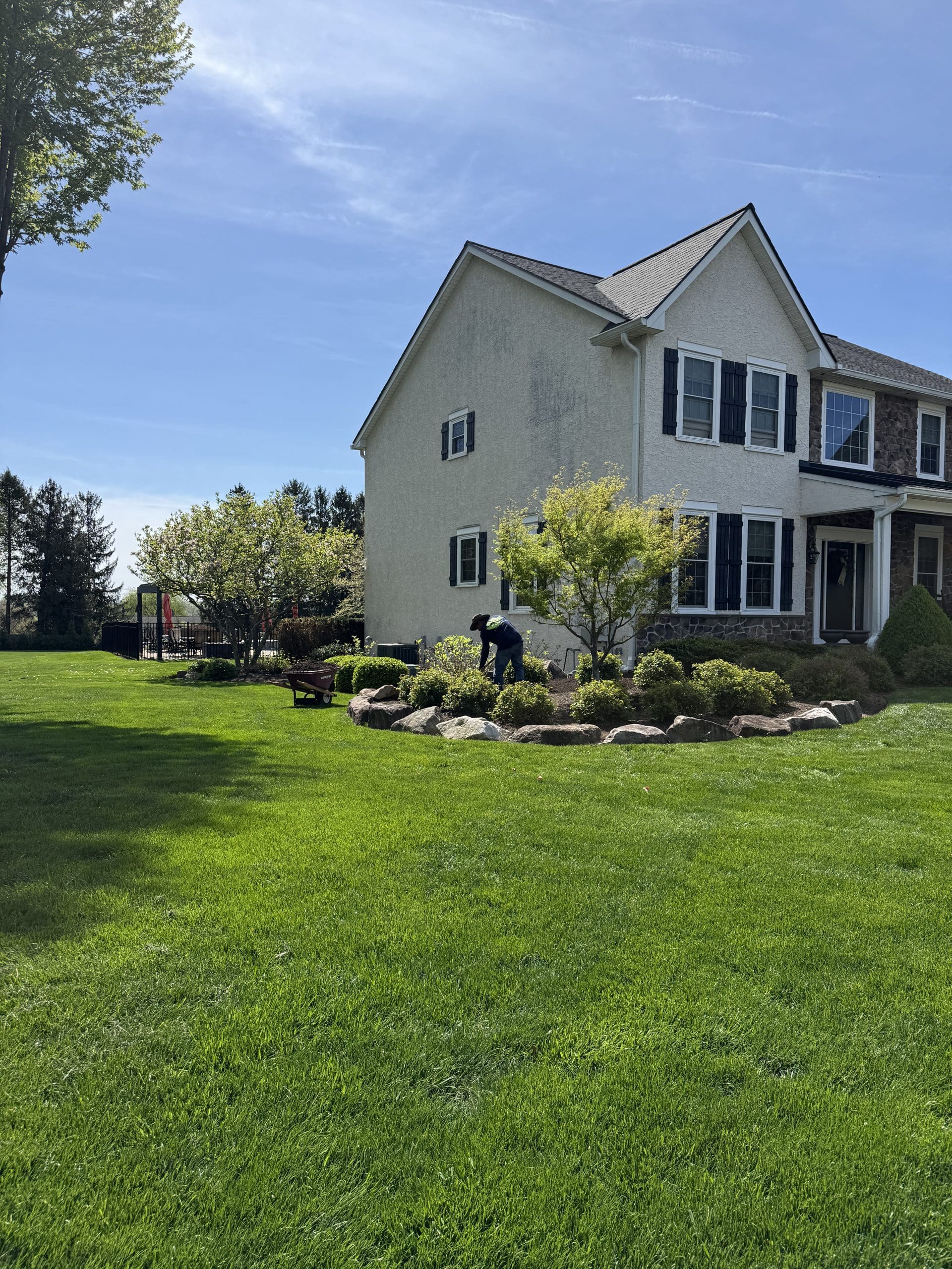 A person is gardening in a flower bed next to a large, light-colored house with black shutters, surrounded by a green lawn and landscaped trees on a sunny day.