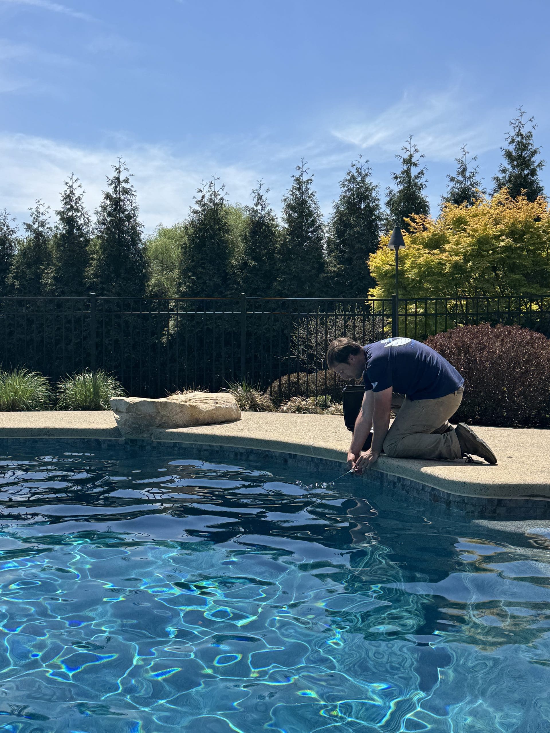 A person kneels at the edge of a clear blue swimming pool, working with their hands near the water, surrounded by greenery and a sunny sky.