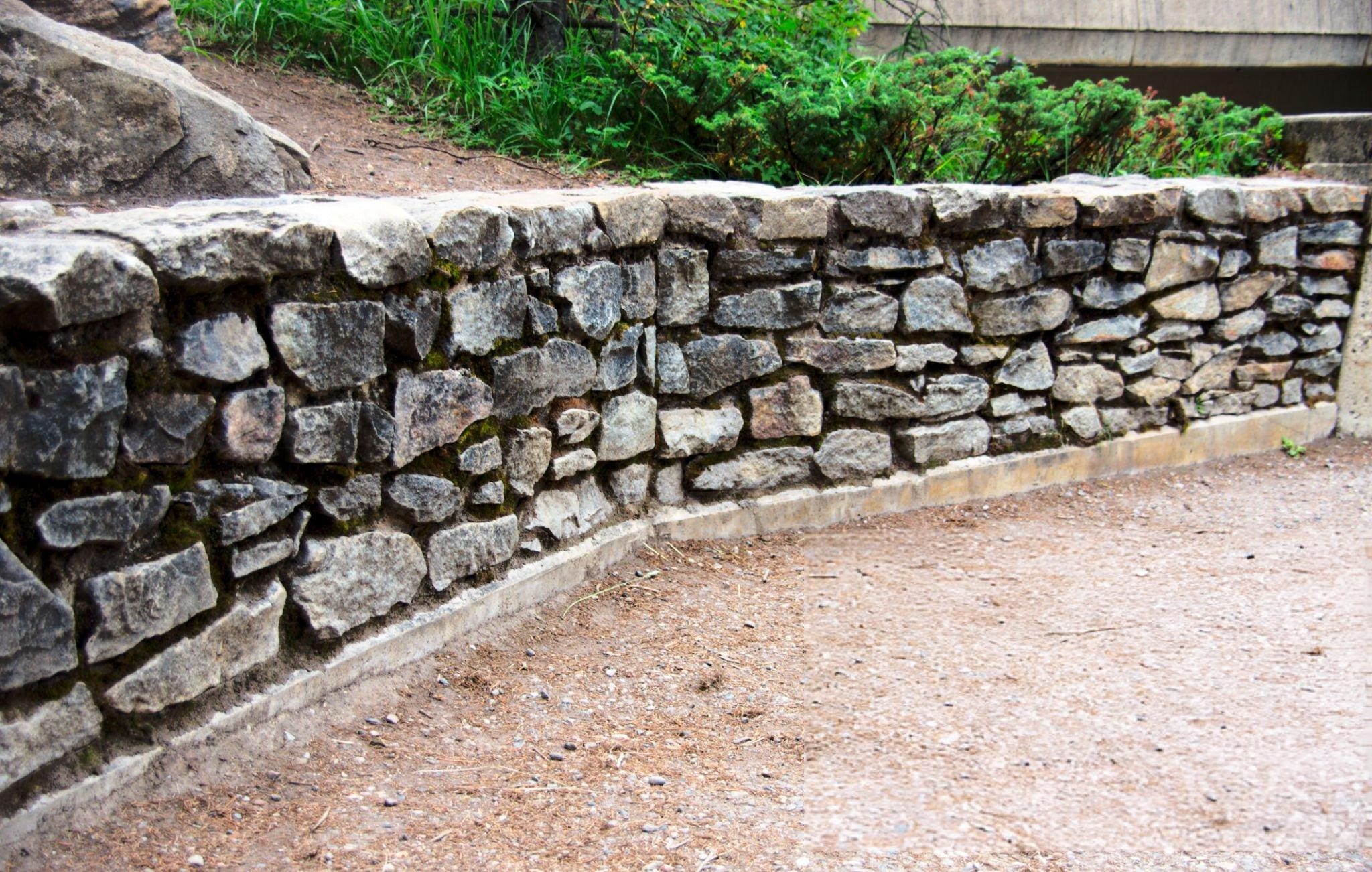 A low stone wall made of irregularly shaped rocks, bordering a dirt path with greenery and shrubs behind it.