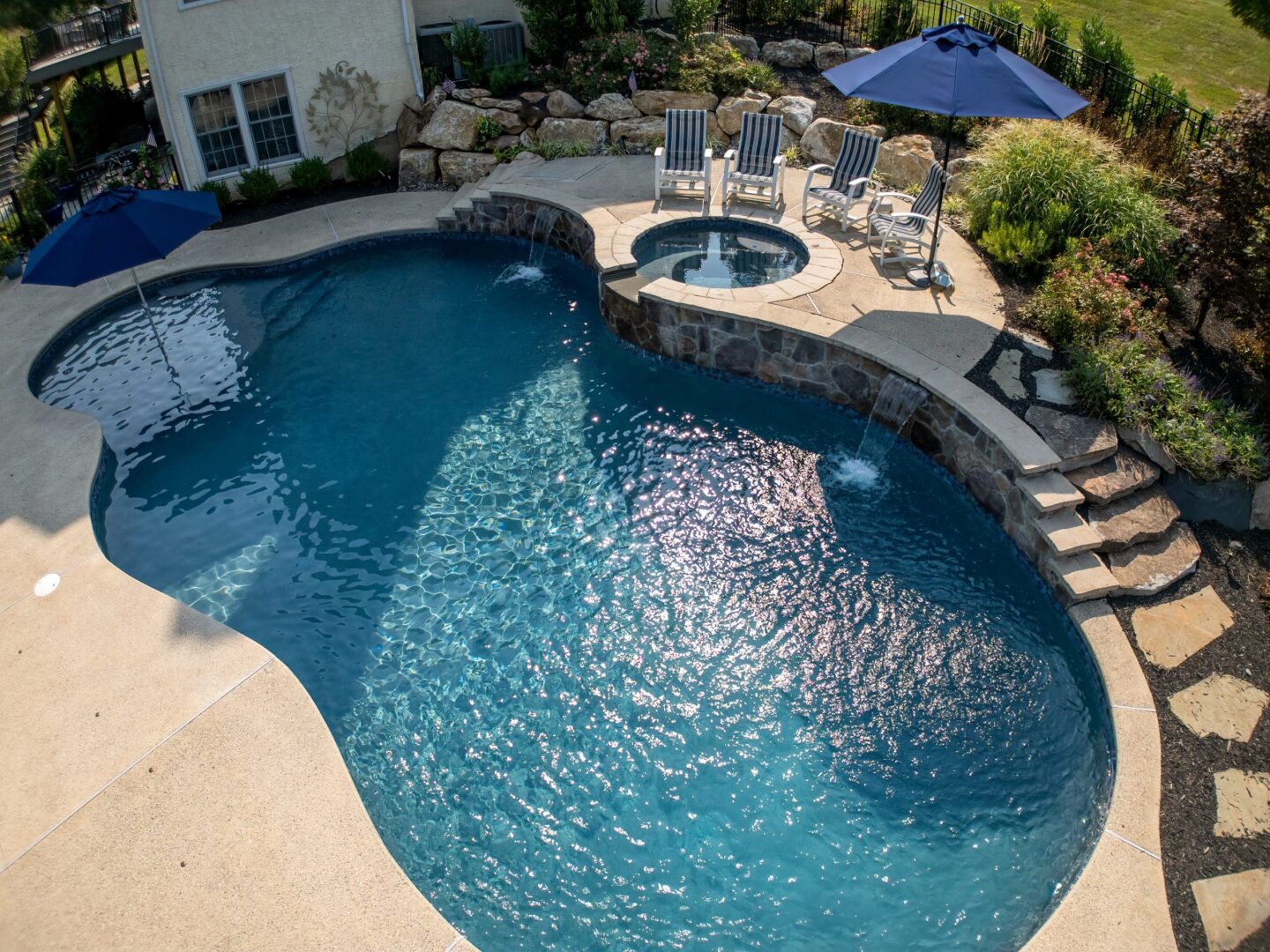 Aerial view of a backyard pool with curved edges, attached circular hot tub, four striped lounge chairs, blue umbrellas, and landscaped garden with large rocks and green plants.