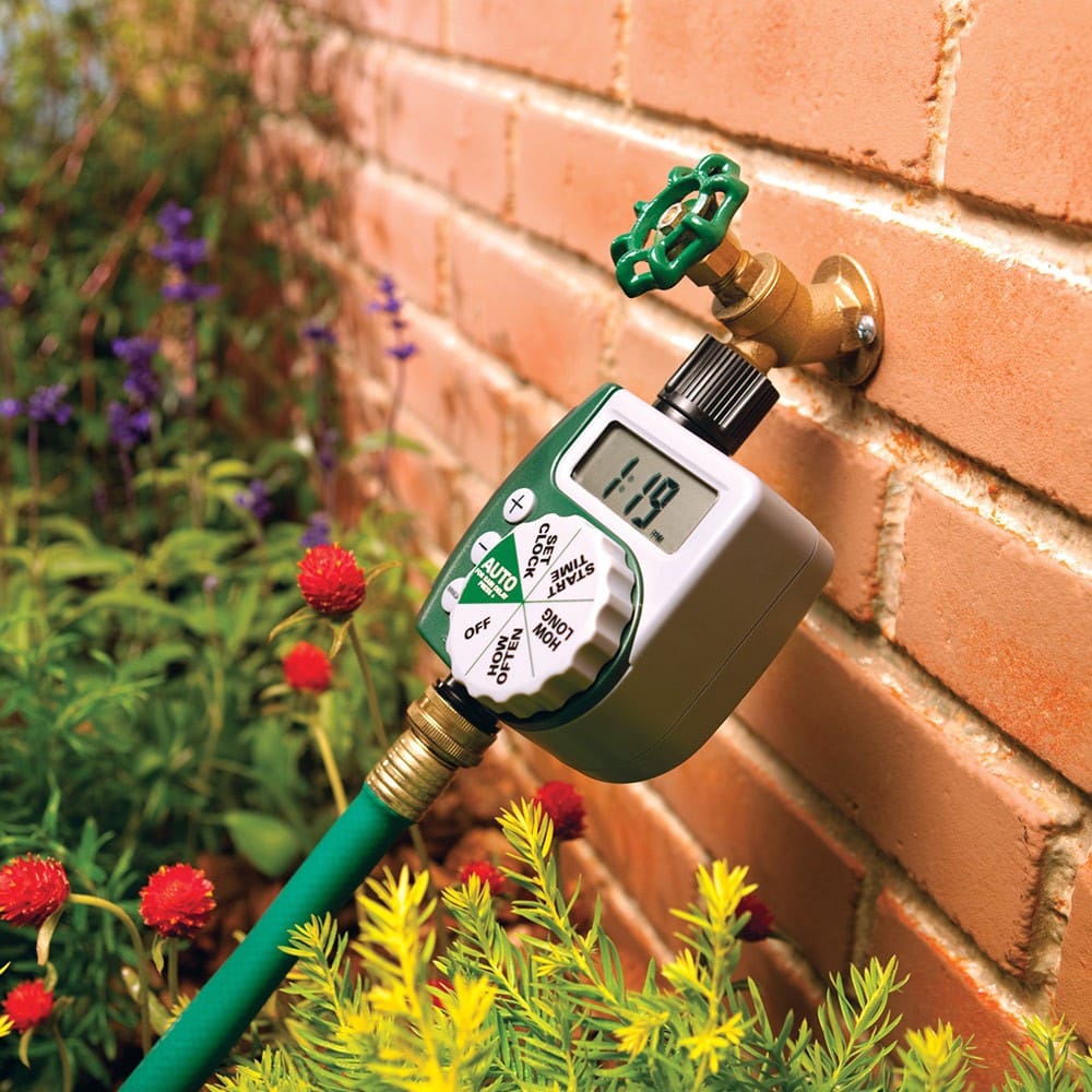 A digital water timer is attached to a garden faucet on a brick wall, with a green hose connected, surrounded by various colorful flowers and plants.