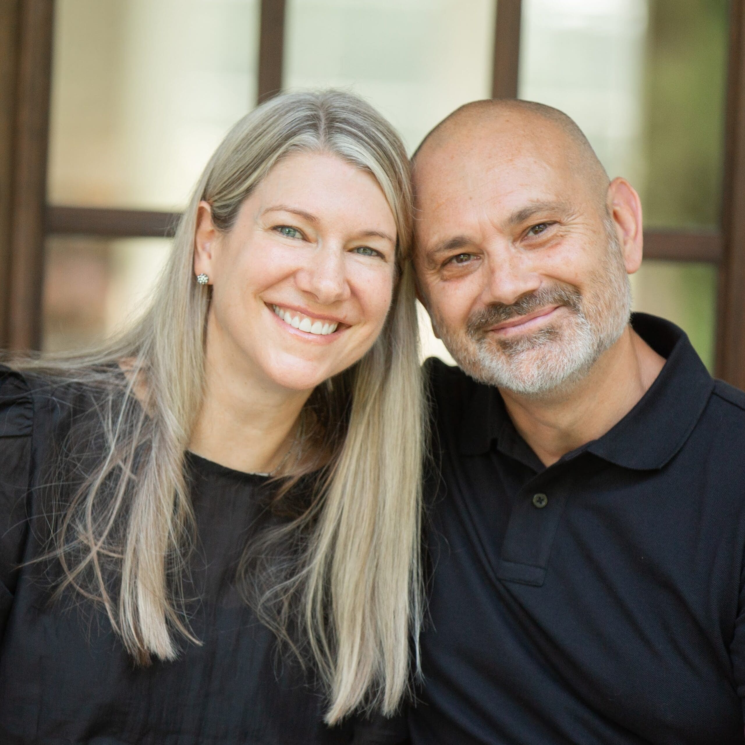 A smiling woman with long blonde hair and a man with a shaved head and beard sit close together, both wearing black shirts, in front of a window.