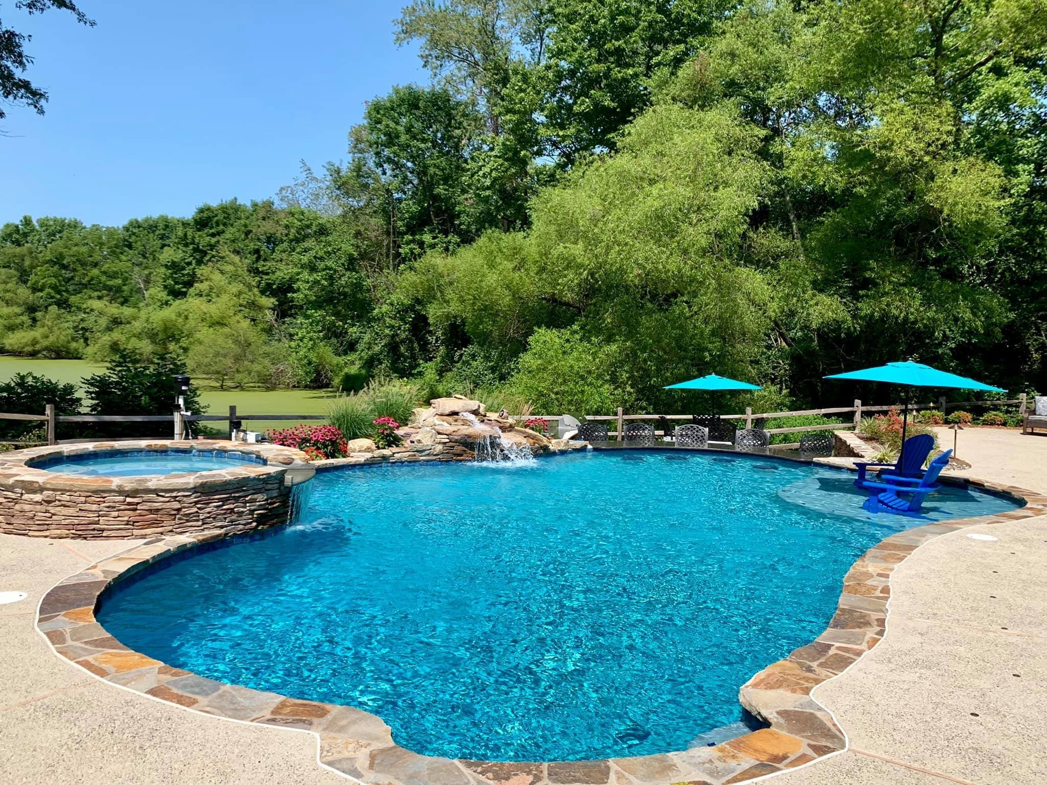 Large outdoor pool with clear blue water, a connected hot tub, stone edging, a small waterfall feature, and two turquoise umbrellas. Surrounded by patio furniture and lush green trees under a blue sky.