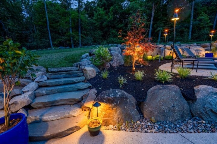 A landscaped Pottstown backyard at dusk with stone steps, garden lights, plantings, lounge chairs, and a circular patio.
