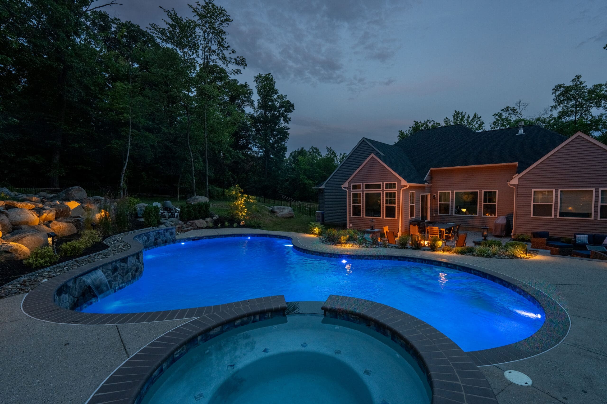 A backyard pool and hot tub are lit up at dusk near a house, surrounded by trees and outdoor seating. The water glows blue, and garden lights highlight landscaping and rocks.