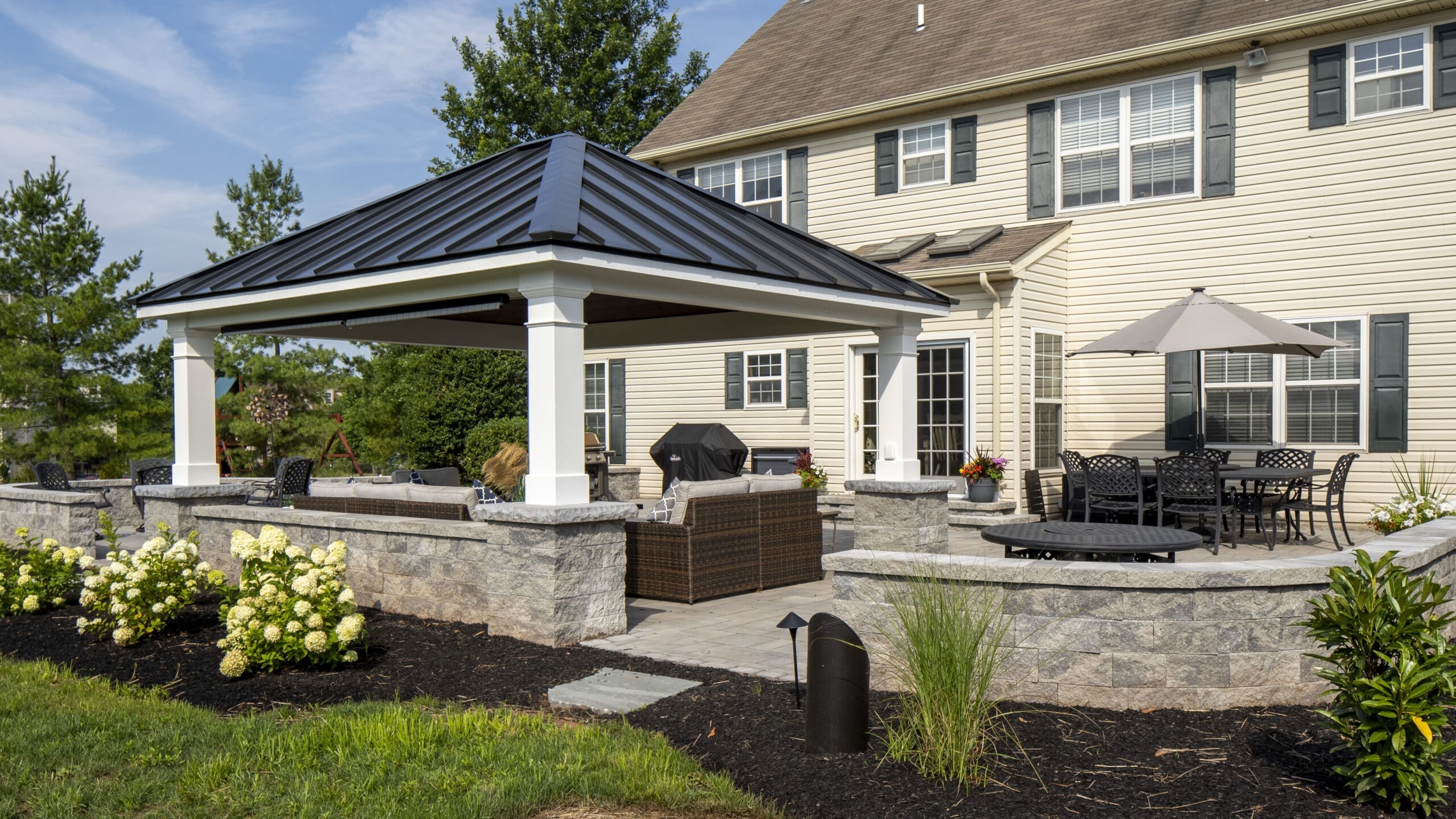A Skippack backyard patio with a white pergola, outdoor seating, dining table, stone walls, and landscaped garden.