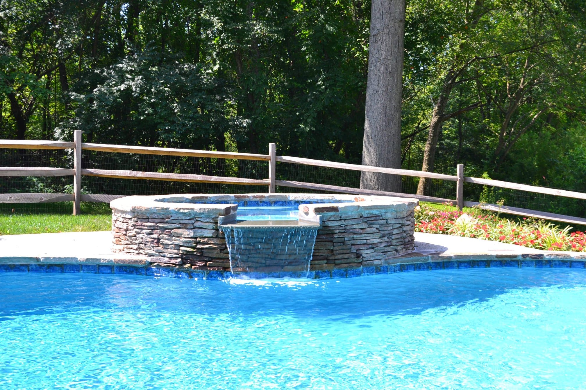 A Southeastern PA backyard pool with a round stone spa and waterfall feature, surrounded by stone decking and trees.