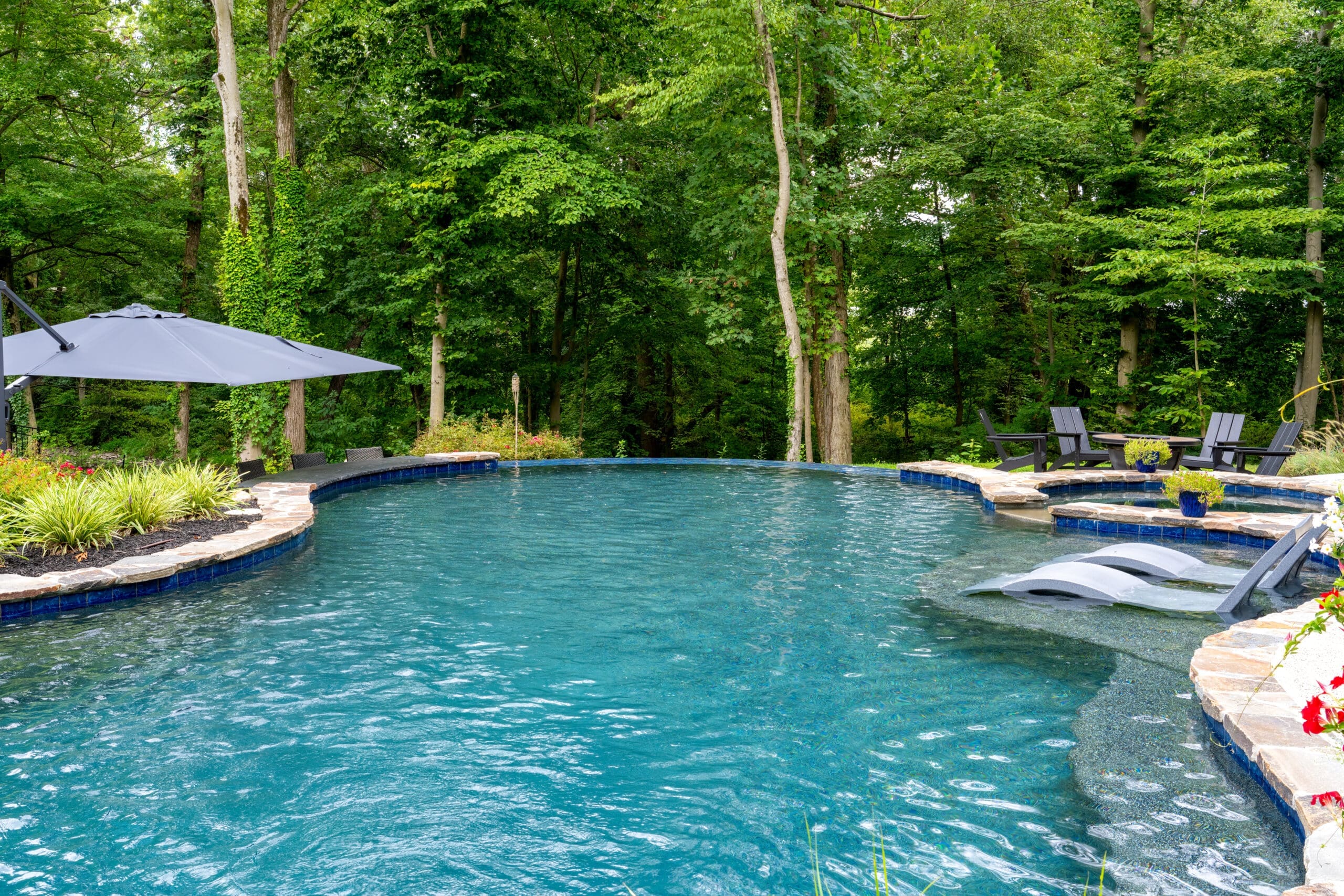 A Southeastern PA backyard pool with a connected spa, waterfall feature, lounge chairs, and lush landscaping.