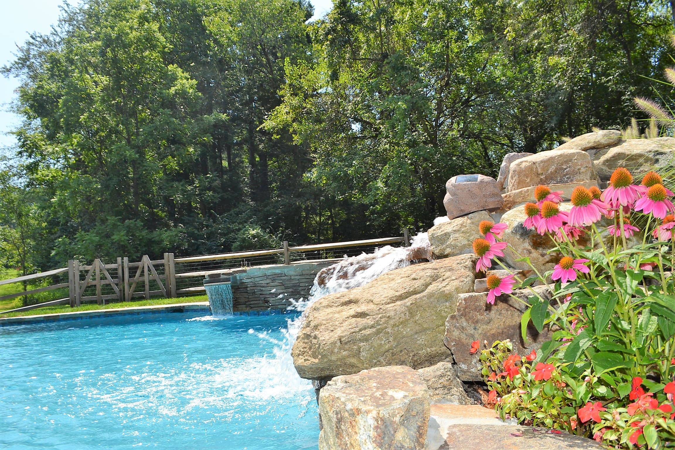 A Southeastern PA swimming pool with a rock waterfall, colorful flowers, and surrounding greenery.