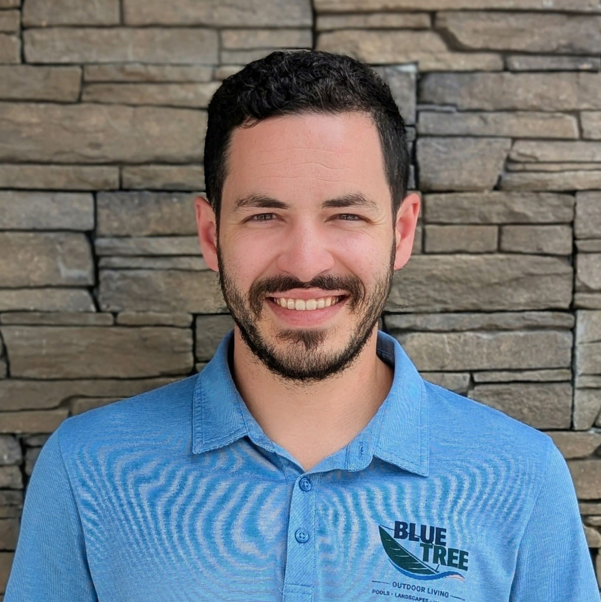 A man with short curly hair and a beard smiles at the camera, wearing a blue polo shirt with a BLUE TREE logo, standing in front of a stone wall.